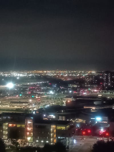 This is an aerial, nighttime photograph of a sprawling urban landscape in Mississauga, Canada. The scene is dominated by a dense concentration of lights emanating from buildings and streets, indicating a developed commercial or residential area. In the foreground, several multi-story buildings with illuminated windows are visible, suggesting office or apartment complexes. Further into the distance, the cityscape stretches out, characterized by a vast expanse of smaller, more scattered lights that blend together, forming a hazy glow against the dark sky. Streets with moving vehicle lights, likely traffic signals and headlights, are discernible in various sections of the image, contributing to the dynamic impression of a city in operation. The overall impression is one of urban activity and illumination under the cover of darkness.