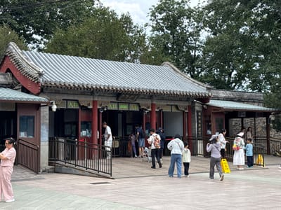 This image captures the New Palace Gate (新建宫门) entrance of the Summer Palace (颐和园) in Haidian District, Beijing, China. The central building features traditional Chinese architecture with a prominent grey corrugated tiled roof, red pillars, and ornate decorative eaves. Multiple entry points are visible, each marked by the green Chinese character "入口" (Rùkǒu), signifying "Entrance."
A group of approximately fifteen individuals, including adults and children, are present on the paved plaza outside the entrance. On the far left, a woman in a light pink short-sleeved top and matching pants is standing, holding and looking at a mobile phone. Near the central entrance, a man in a white short-sleeved shirt and checkered pants stands at a turnstile. Other individuals are distributed across the plaza, with several walking towards or through the entrance points. One man is observed pushing a stroller. A security camera is mounted above one of the central entryways.
The building complex includes various sections. The central structure has red pillars and ornate detailing, while a section on the right features a stone-patterned wall and a smaller, green-tiled roof. Two yellow A-frame caution signs are placed on the ground near the right side of the entrance. The ground consists of light-colored, patterned paving stones. In the background, numerous large, green trees are visible beneath a partially cloudy sky. Overhead utility wires traverse the upper left portion of the image.