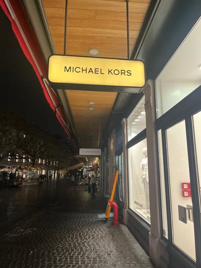 This media file captures an urban street scene in Genève, Switzerland, at night or dusk. The street is lined with commercial buildings featuring large display windows and illuminated signs.
The most prominent feature in the foreground is a bright yellow, vertically oriented sign displaying "MICHAEL KORS" in black capital letters. This sign is positioned next to a building facade with wooden paneling and several recessed circular lights. Below the sign and along the building's base is a stretch of red material, possibly a carpet or awning, extending into the dark foreground.
Above the "MICHAEL KORS" sign are storefront windows, reflecting the opposite side of the street. Partial text, "MICHAEL KORS'S COLLECTION," is visible within one of these window displays. Further down the cobblestone street, which appears wet and reflective from recent rain, a distinct red pipe structure with a yellow angled arm is visible.
In the background, the street recedes into the distance, lined with more illuminated shops and what appear to be bare trees, suggesting a winter season. Decorative lights are strung across the street or on buildings, adding to the evening ambiance. A few blurry figures can be seen walking in the distance, indicating pedestrian activity. Other inverted signs are faintly visible, one seemingly reading "ABENDELEGANT" and another "WINTER." The overall scene portrays a damp, active commercial street in a European city after dark.