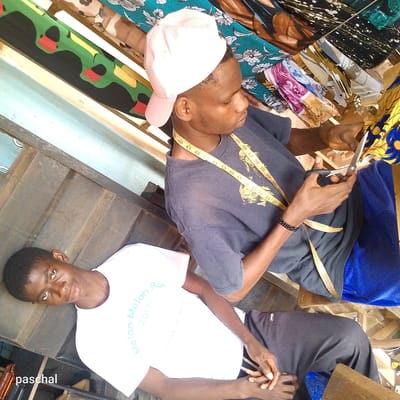 The image shows two men in what appears to be a tailor's shop in Nsugpe, Nigeria. One man is seated, wearing a gray t-shirt with a measuring tape around his neck, and a light pink baseball cap. He is holding scissors and cutting a patterned fabric. The other man is reclining, wearing a white t-shirt with the words "Melon Melon Run 2017" printed on it. The environment includes various fabrics and materials indicative of a tailoring business. The backdrop shows different colored fabrics, suggesting they are materials for clothing. The setting seems to be a modest and possibly cramped workspace.