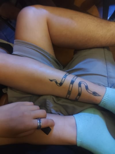The image shows a close-up view of a person's legs and a snake tattoo on their forearm. The person is wearing grey shorts and light blue socks. A silver ring with a chain pattern is visible on the person's finger. The tattoo is black and depicts a stylized snake wrapping around the forearm, with segmented bands along its body. The environment appears to be indoors, possibly a home, with the background out of focus, suggesting a casual setting. There is no information in the image that indicates the location in the city of Lozenets, Bulgaria.