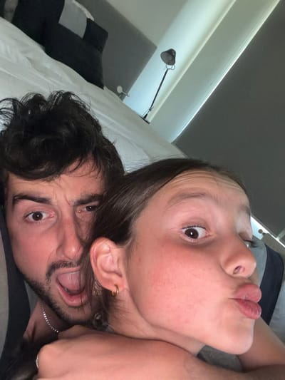Two people are close together in a selfie, lying in what appears to be a bed. The person on top is a fair-skinned woman with dark hair and a gold hoop earring. She is pouting her lips toward the camera. The person below her is a man with dark hair and a beard, making a funny face with an open mouth and wide eyes. Pillows and a lamp are visible in the background. The photo is taken at an angle, making the subjects appear slightly distorted.