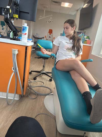 A young girl sits in a teal dental chair in a brightly lit dentist's office. She is wearing a white t-shirt with a cat graphic and the word "PURRFE" on it, black shorts, black socks, and grey shoes. Her legs are crossed. She is holding a bright yellow dental tool in her right hand. The dental office has orange cabinets, a black monitor on an articulating arm, and other dental equipment. There are stuffed animals and decorative boxes on a shelf in the background. The floor is light-colored wood. The image was taken in Chesapeake, United States of America.