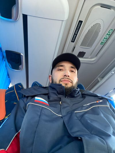 A selfie of a man in Koskolovo, Russia, on what appears to be an airplane. The man is wearing a black baseball cap and a navy jacket with the Russian flag on the sleeve and the word "AVIACOMPANY" in Cyrillic script. The plane's interior is white with black trim. A sign in Russian that translates to "Emergency Exit" is visible above the man's head. The seat covers are blue and gold.