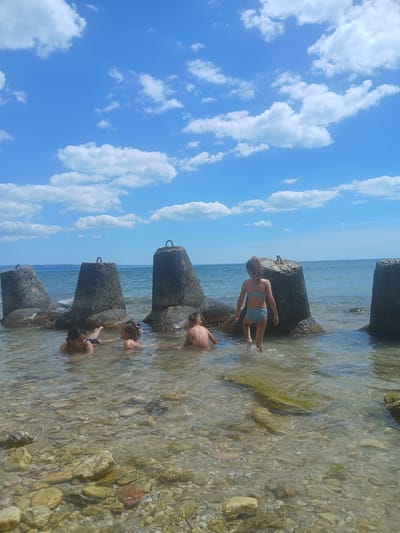 In a coastal scene in Osenovo, Bulgaria, several children are playing in the shallow water near concrete tetrapods. The sky is a brilliant blue with scattered white clouds, suggesting a clear, sunny day, likely during daytime hours. The water is clear and shallow, revealing the rocky seabed below.

One child, wearing a blue bikini, is standing on a tetrapod and appears to be reaching out. Another child, a boy in swim trunks, is partially submerged in the water. Further down, two more children are also playing in the water. Their interactions suggest a playful and relaxed atmosphere, typical of a summer day at the beach. The tetrapods, used as breakwaters, create a somewhat rugged and industrial element within the natural coastal landscape. The overall impression is one of children enjoying a refreshing dip in the sea on a warm, bright day. There is no visible text in the image.