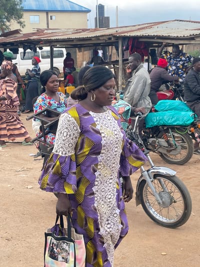 The image depicts a bustling outdoor market scene in Wang, Nigeria, likely during daytime. Several people are present, with a focus on a woman in the foreground wearing a vibrantly patterned purple and yellow dress with white lace detailing. She is looking downwards and holding a tote bag with a colorful tie-dye pattern featuring images. Behind her, another woman in a floral print dress is partially visible, also looking towards the right.
In the background, a motorcycle is prominent, with a turquoise tarp draped over its handlebars. Several other individuals are seen around the motorcycle and a rustic, open-air structure with a corrugated metal roof. One person in a blue and black striped shirt and a red cap is leaning towards the motorcycle. Further back, a white van is parked, and more people are visible, suggesting a busy commercial area. The ground is sandy and dusty, characteristic of an outdoor market. The overall atmosphere appears lively and active, with people engaged in daily activities within the market. No specific time of day or weather conditions are discernible beyond the general brightness of daylight.