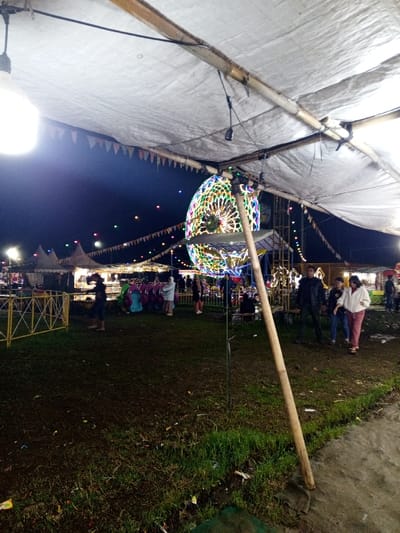 Under a makeshift tent lit by hanging bulbs, an outdoor fair bustles at night. People stroll through the grassy area, with a brightly lit Ferris wheel in the background. Strings of colored lights and pennant banners add to the festive atmosphere. A man and two younger people stand together near the right side of the scene.