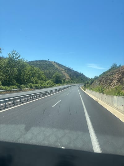 The image depicts a view from a vehicle traveling on a highway near Avra, Greece. The highway has multiple lanes separated by white lines. On the left side of the road, there is a metal guardrail and a steep, tree-covered hillside. The right side of the highway features a concrete barrier and a rocky embankment with sparse vegetation. The sky is clear and blue, with a few wispy clouds visible. The terrain appears hilly and somewhat arid, with evidence of past forest fires on some of the slopes, indicated by bare trees and scorched earth. One car is visible in the distance on the highway. The time of day appears to be daytime, likely mid-morning or early afternoon, given the bright sunlight and clear sky.