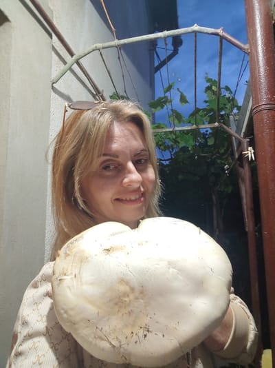 A woman with blonde hair and sunglasses pushed up on her head smiles at the camera while holding a large, white mushroom in front of her. She is wearing a patterned sweater. Behind her, a vine-covered trellis is visible against a twilight sky. The scene appears to be an outdoor area, possibly a backyard or garden, with concrete walls and a brown pipe structure to the right. The location is specified as Alushta, Ukraine.