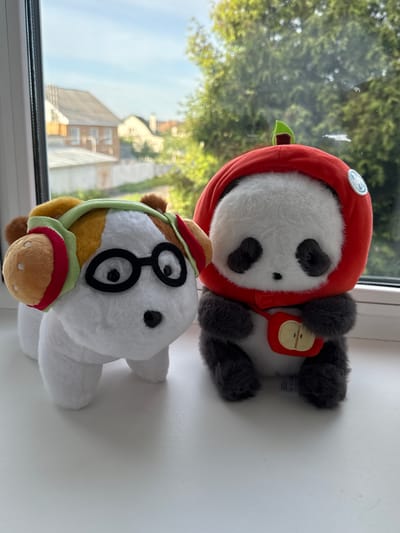 Two plush toys sit on a white windowsill. The toy in the foreground is a white horse wearing black-rimmed glasses and a brown and red-trimmed hat and halter. Behind the horse is a panda bear plush wearing a red hood with a green apple stem detail and a red apple-shaped backpack. The background, visible through the window, features blurry foliage and buildings, suggesting an outdoor scene in Severnyi, Russia.