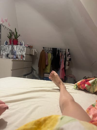 A person is lying on a bed in a room with a slanted ceiling. The room contains a garment rack filled with clothing and a set of drawers. There is a plant on a shelf near the drawers. The location is presumed to be in Saarbrücken, Germany.