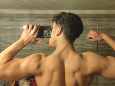A person faces away from the camera, holding a smartphone up to their face. The individual flexes both arms to display muscular development in the biceps, triceps, shoulders, and back. A tiled wall background shows a horizontal pattern. Miscellaneous objects are partially visible in the lower left corner.