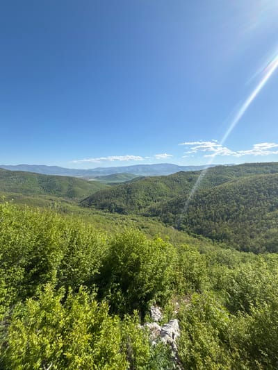The image depicts a mountainous landscape under a clear blue sky with scattered clouds. The scene is dominated by green trees and vegetation covering the slopes. In the distance, a valley and distant hills are visible. Sunlight streaks across the sky. The location is likely near Lipnitsa, Bulgaria, based on the context. No people are visible.