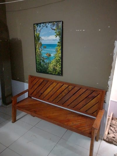 The image shows a wooden bench in front of a wall. A framed picture of a tropical beach scene hangs on the wall above the bench. The bench is made of polished wood and has a slatted backrest. The wall is painted a neutral color and appears to be slightly damaged in some areas. The floor is tiled. The lighting suggests it is indoors, possibly in a residential or commercial setting. No people are visible in the image. The specific location in Cuiabá, Brazil, cannot be determined from the image alone.