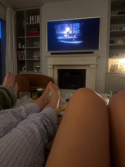 This image captures a cozy living room scene, possibly in Rueil-Malmaison, France, where two individuals are enjoying a movie night. The focus is on their legs and feet, suggesting a relaxed and intimate atmosphere. The television screen displays a scene from a movie, with a fireplace below it. Bookshelves filled with books and decorative items line the walls, adding to the homey ambiance. There are also drinks and snacks on a coffee table, indicating a comfortable movie-watching setup. The lighting is dim, creating a warm and inviting mood. The overall impression is one of leisure and shared enjoyment in a well-appointed home.