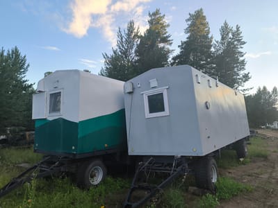 The image shows two mobile units parked on a dirt and grass lot. The unit on the left is white with a green stripe. It has a window on the side and sits on a trailer with large wheels. The unit on the right is gray, also with a window on the side, and is on a similar trailer. Both trailers have towing hitches. Behind the units, there are numerous pine trees. The sky above is blue with scattered clouds, some of which are lit by sunlight. The scene appears to be in a rural or semi-industrial area, possibly on the outskirts of a settlement like Гремучий, Russia. There are no people visible in the image. The overall impression is one of storage or temporary housing in an outdoor setting.