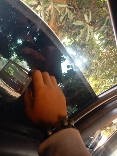 The image depicts an interior view from a vehicle located in Unguwar Gaya, Nigeria, looking outwards through a partially lowered window. In the foreground, a light brown hand, adorned with a multi-beaded bracelet containing dark spherical and rectangular beads, is visible. The individual is wearing a light grey long-sleeved garment with a ribbed cuff. Sections of the vehicle's dark interior, including the window frame and door panel, are observable. Outside the window, dense green foliage from trees is prominent, with bright sunlight penetrating the leaves on the right side of the frame, generating a distinct sun flare. In the lower-left background, a light-colored structural element, potentially a wall, and a wooden frame structure are discernible above light-colored ground. The scene is illuminated by natural daylight.