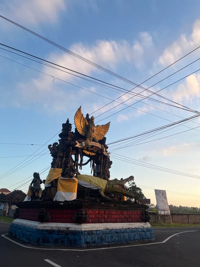 The image depicts a monument in the city of Mendoyo, Indonesia, under a bright blue sky with scattered clouds and a network of overhead power lines. The monument is a multi-tiered structure with intricate carvings, prominently featuring a large golden eagle with outstretched wings at its apex, bearing the inscription "BHINNEKA TUNGGAL IKA". Below the eagle, various figures and mythological creatures are depicted, including a fierce-looking crocodile with its mouth agape. The monument's base is adorned with red and blue colored blocks. Yellow and white cloths are draped around parts of the structure, possibly as offerings or decoration. To the left of the monument, traditional Indonesian houses can be seen. To the right, a large banner is displayed on a wooden stand, and a brick wall lines the background. The monument is situated at a road intersection, with asphalt roads visible in the foreground. The overall scene suggests a cultural or religious landmark in a semi-rural setting.