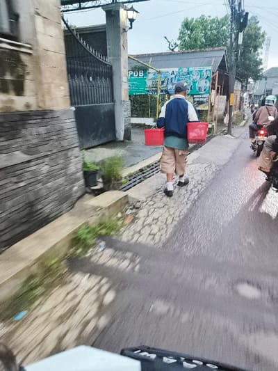 The image was taken from the perspective of someone on a motorcycle, facing forward. The location is Cimenyan, Indonesia, coordinates -6.886 latitude and 107.653 longitude. The scene is a narrow street with a grey concrete surface. A man is walking on the left side of the street, carrying two red baskets in his hands. He is wearing a blue jacket, beige shorts, and a blue cap. The man is walking away from the camera. The motorcycle appears to be moving forward, creating a blurry motion effect in the foreground and background. There is a white building on the left side of the frame, and the image is taken on a cloudy day.