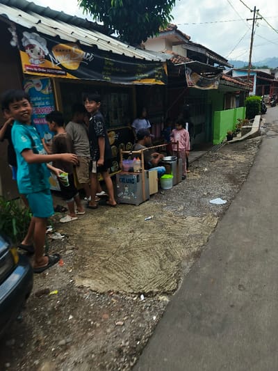 The image shows a street scene in Cimenyan, Indonesia, located at coordinates -6.885, 107.654. A group of people are gathered in front of a small storefront with a sign that reads "Dapur Saci". There are several men and women standing around a small table where a man is seated, apparently preparing food. The storefront is adorned with a yellow awning and has a sign advertising "smoothies" on the wall. There are a few potted plants on a small wall behind the group, and an alleyway leads to the left, toward a paved street. The pavement is cracked and worn. To the right of the storefront, there is a small house with a green wall. A motorcycle can be seen in the background, and there are several power lines in the sky. The people are standing in a line, some of them looking towards the camera. The person in the forefront is wearing a blue and green t-shirt and shorts. They are standing with their feet on the ground and their hands in front of them. The people in the background are blurred, but they appear to be moving around the table and the storefront. The scene appears to be a typical street scene in a small town in Indonesia.
