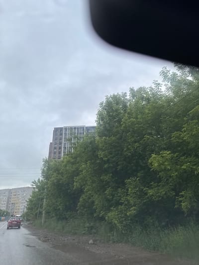 The scene depicts a street view in the city of Кондратово, Russia, under an overcast, light grey sky. The image appears to be captured from inside a vehicle, with a large, dark, out-of-focus curved object, consistent with a dashboard or rearview mirror, partially obscuring the upper right quadrant of the frame.
In the background, two multi-story buildings are visible. To the far left, an older, light-colored residential apartment block with multiple windows and balconies is present. To its right, partially obscured by trees, stands a taller, more contemporary building featuring a two-tone facade: darker, brick-like sections on the left and lighter sections with prominent vertical windows on the right.
The midground is dominated by a dense line of mature, deciduous green trees and shrubs, extending horizontally across the image and partially concealing the newer building. Several thin, dark utility lines are visible traversing through this foliage.
In the foreground, a wet asphalt road with numerous visible potholes and patches of standing water extends from the bottom left towards the center-right. A red sedan is prominently featured on this road, driving away from the viewer, with a partially visible white vehicle behind it. To the right of the road, a dirt and gravel shoulder, also wet and muddy, is present, bordered by tall green grass and weeds. A utility pole and a yellow traffic light are visible on the left side of the road, near the red car. No individuals are distinctly visible.