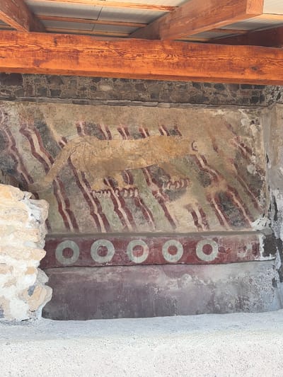 The image shows a section of an ancient mural, likely within the archeological site of Teotihuacan, near San Francisco Mazapa, Mexico. The mural depicts a stylized figure in a faded, ochre color, with details suggesting possible ceremonial clothing or adornments. The figure stands against a background of alternating red and darker colored horizontal stripes. To the right of the figure is a vertical band with five concentric circles, also in a faded color. The mural appears to be deteriorating, with signs of age and damage to the paint and plaster. Modern wooden beams are visible on the left side of the image, indicating current structural support or preservation efforts. The mural is flanked by sections of a gray stone wall on the left and a newer, white-colored wall on the right. Debris is piled at the base of the mural.