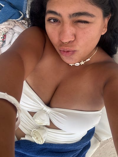 A woman is lying down on what appears to be a beach, as suggested by the presence of sand and beach towels. She is wearing a white swimsuit with decorative floral knots and a seashell necklace. She has dark hair, dark eyebrows, and a gold nose ring. One eye is open and looking directly at the camera while the other is closed in a wink. A blue towel and a crocheted fabric are visible beneath her. The photo is taken at a close-up angle, offering a clear view of the woman’s face and upper body. The location is assumed to be Playa del Carmen, Mexico.