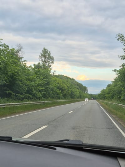 The image depicts a view from inside a vehicle traveling on a two-lane highway lined with dense green trees and vegetation. The road surface is asphalt with white lane markings. In the distance, several vehicles are visible on the road, including a white van and at least two cars. The sky is overcast with clouds, but there are hints of lighter sky and possibly a sunset or sunrise in the far distance, with a patch of light orange and blue clouds. The scene appears to be a rural or semi-rural setting, possibly on the outskirts of Niva, Russia. The perspective is from the front passenger seat or driver's view, looking through the windshield, which is partially visible at the bottom of the frame.