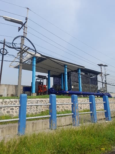 The image captures an outdoor industrial or utility facility under a clear, bright sky, likely in Kecamatan Gempol, Indonesia. The main structure, situated on the right side of the frame, features a dark-colored roof supported by prominent blue vertical beams. Beneath the roof, various pieces of machinery are visible, characterized by their orange-red components and intricate blue piping systems, suggesting a function related to water management, pumping, or a similar industrial process.
In the mid-ground, a horizontal barrier or fence runs across the scene. It is constructed from multiple parallel blue concrete or painted metal bars, supported by vertical grey metal posts. This barrier rests on a textured wall made of stones or concrete. To the right of the barrier, a patch of vibrant green grass is visible.
The left side of the image is dominated by several utility poles extending upwards, supporting a complex network of black power lines that crisscross against the light blue sky. Mounted on these poles are various electrical components, including what appears to be a solar panel or antenna structure positioned higher up.
The scene is illuminated by natural daylight, indicating clear weather. There are no people present, and no specific activity is taking place, presenting a static view of functional infrastructure. No visible text is legible in the image.