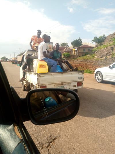 In Kuru, Nigeria, on a bright, overcast day, five individuals are being transported in the open bed of a white utility truck. The truck is traveling on a paved road, possibly part of a highway, with a white car visible in the right lane. The truck is loaded with various items, including a large yellow plastic container with a red lid, bags of produce, and a blue bag labeled "PRAYER."
The individuals in the truck are seated and standing. Two men are seated in the front of the truck bed, one wearing a green shirt and dark pants, the other in a white shirt and blue pants. Behind them, three more men are positioned, one standing and wearing an orange and black patterned shirt, and two others seated behind him. The man standing appears to be looking towards the camera.
The environment includes a rocky, grassy hillside in the background with some buildings and trees. Power lines are visible stretching across the sky. The road surface shows signs of wear and tear, with cracks and patches. A side-view mirror of a vehicle, presumably the one from which the photo was taken, is prominently featured in the foreground, reflecting a blue container and part of the truck's interior. The overall scene suggests a common mode of transportation in the region, possibly for labor or goods.