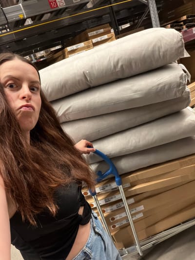 A person with long brown hair, wearing a black top and blue jeans, is in what appears to be a retail or warehouse setting. They are holding the handle of a blue cart containing rolls of fabric or material, and cardboard boxes. The location is possibly a store in Kaiserslautern, Germany.