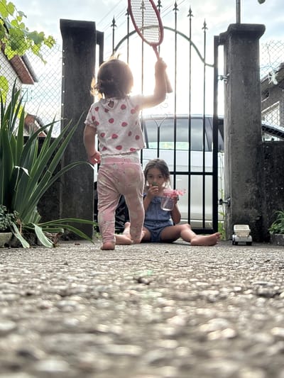 In Oreshak, Bulgaria, two young children are playing outdoors near a residential gate. A toddler, seen from behind, stands holding a red badminton racket raised above her head. She wears a light-colored shirt with a strawberry pattern and light pink pants with a fruit motif. To her right and slightly in front, a younger child sits on the ground, wearing a blue and white striped romper. This child holds a pink sippy cup and appears to be eating something green. A small white toy truck is visible on the ground to the right of the seated child. The scene unfolds on a textured concrete ground, with a metal gate and stone pillars forming a backdrop. Lush green foliage is present to the left of the frame, and a fence is visible behind it. The sun is setting or rising, casting a bright light from behind the gate, creating a silhouette effect.