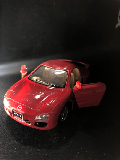 A red toy Mazda RX-7 car is positioned on a black textured surface. The car is slightly angled, with the driver's side door and the passenger side door ajar. The front of the car displays the Mazda logo and "RX-7" on a license plate. The car's headlights are visible, and one wheel is partially obscured. The background is dark and out of focus, suggesting a dimly lit environment. The scene does not contain any people or indicate any specific location within the city of Yelizovo, Russia.