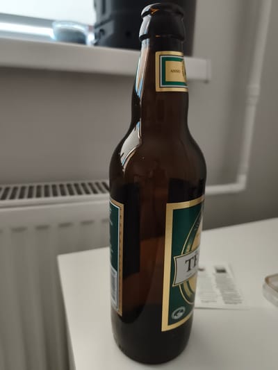 A brown glass bottle of beer, with a label featuring green and gold accents, stands on a white surface. The bottle is positioned to the right of the frame, with its neck tilted slightly to the left. In the background, a white radiator is visible, and further back, a light-colored wall and a window are present. A small object, possibly a lid or cap, is partially visible on the far right of the white surface. The scene appears to be indoors, likely in a domestic or office setting. There are no people in the image.