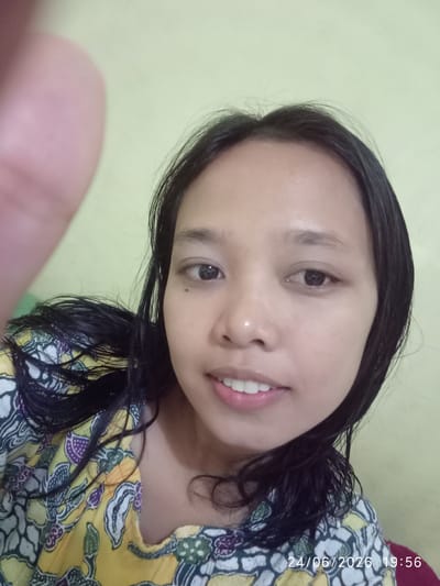 A young woman is shown in a close-up selfie, facing the camera directly. She has long, dark, slightly wet hair framing her face. Her eyes are dark brown, and she has a small beauty mark below her left eye. She is smiling faintly, revealing white teeth. She is wearing a yellow garment with a colorful batik-style pattern of abstract shapes and floral motifs in white, purple, and green. The background is a plain, light green wall. In the lower right corner of the image, a timestamp is visible: "24/06/2026 19:56". The overall impression is casual and personal, likely taken indoors.
