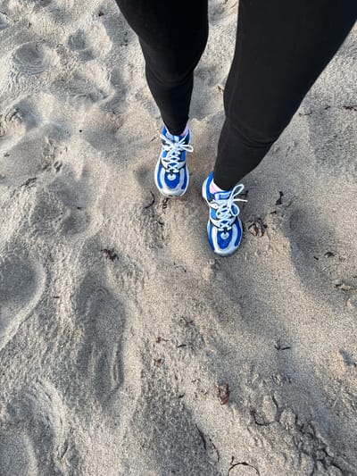 A low-angle shot captures a person's legs wearing black leggings and blue and white athletic shoes as they walk on a sandy surface. The sand is textured with light and shadow, suggesting uneven terrain. Small dark pieces, possibly seaweed, are scattered across the sand. The context of Teriberka, Russia, is not visually discernible from the image itself.