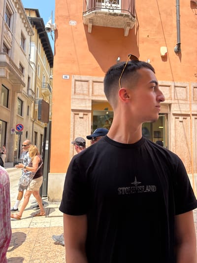 The image shows a young man in a black Stone Island T-shirt standing on a sidewalk in Verona, Italy. He is looking up, with sunglasses perched on his head. Behind him is an orange building with visible architectural details, including a "16" sign. In the background, other people can be seen walking on the sidewalk, and there are additional buildings lining the street. A "no parking" sign is also visible. The scene appears to be taken during daylight hours.