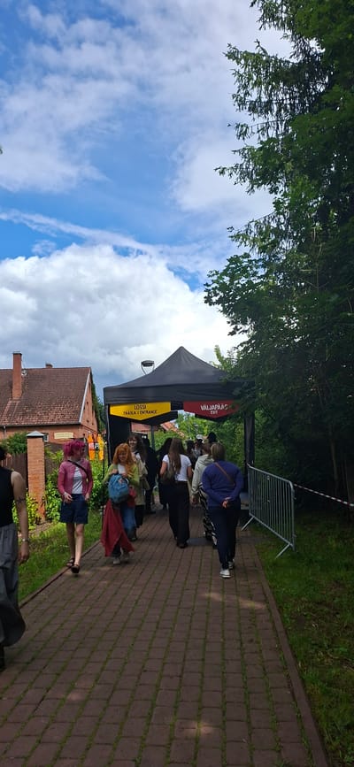 A group of people are walking on a paved walkway towards a black tent. The tent has yellow and red signage that reads "LOSSI PÄÄSLÄ / ENTRANCE" and "VÄLJAPÄÄS EXIT." To the left of the walkway is a brick building with a red roof and a brick fence. To the right of the walkway is a metal fence and green grass. The sky is blue with white clouds. The scene appears to be an outdoor event in Viljandi, Estonia.