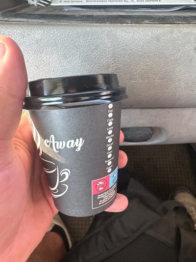 A person's hand holds a black paper coffee cup with a black lid. The cup has white text that says "Away" in cursive. Below "Away" there is an illustration of a coffee cup. There is a list of coffee options on the side of the cup: "Flat", "Capp", "Latte", "Mocha", "Decaf", "Choc", "Sugar.0", and "Sugar". The bottom of the cup has an emblem that says "PLASTIC IN PROD". The background appears to be the interior of a vehicle, possibly a car or bus, with grey textured plastic. A black bag and a shoe are visible in the background. The location is identified as Livadia, Cyprus, based on a sticker at the top of the image which reads "mot larnaca BIOTEΧΝΙΚΗ ΠΕΡΙΟΧΗ No. 10, 6025 ΛΑΡΝΑΚΑ".