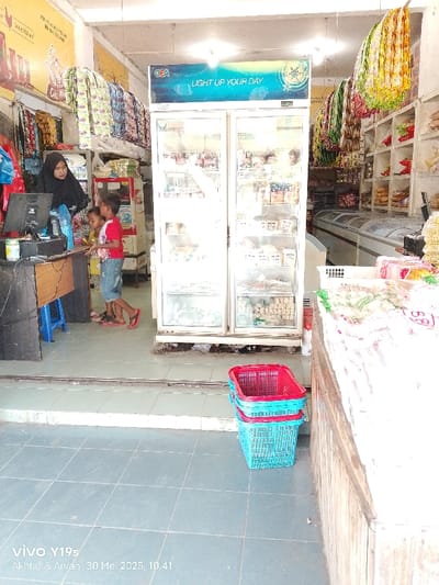 This media file depicts the interior of a small shop or minimarket, likely located in Kecamatan Haurgeulis, Indonesia, during the late morning, specifically at 10:41 AM on May 30, 2025, according to the visible watermark.
The scene features a woman, clad in a black hijab and dark clothing, standing behind a wooden counter on the left. She appears to be engaged with a computer monitor and keyboard, suggesting she is managing transactions or inventory. Two young children are positioned next to her, looking at the counter. One child wears a red t-shirt and blue shorts, while the other is in a yellow and white striped t-shirt. Both children are wearing sandals or are barefoot. A blue plastic stool is partially visible under the counter.
Central to the image are two large, white, double-door glass refrigerators, showcasing various packaged items inside. A blue banner above them prominently displays the text "CEA LIGHT UP YOUR DAY". To the right of these refrigerators, the shop extends further back, revealing shelves stocked with numerous products and a long, white horizontal freezer. Various packaged goods, possibly snacks or small items, hang from display racks or lines near the ceiling and walls.
In the foreground on the right, three plastic shopping baskets are stacked: a red one on top of two blue ones. The floor throughout the shop is covered with grey tiles. The overall setting portrays a typical day at a local store, with the woman attending to duties while the children are nearby. The camera model "vivo Y19s" and names "Akhtar & Arvan" are visible watermarks at the bottom left.