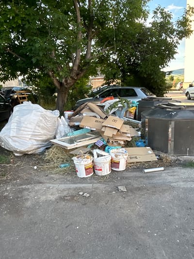 In Kostinbrod, Bulgaria, this image depicts an outdoor scene on a paved surface, likely a street or alley, with a large pile of accumulated waste. The waste consists of various items, including large white plastic bags filled with debris, numerous flattened cardboard boxes, some of which are labeled "IKEA," a discarded window frame with glass, and several white buckets. Some of the buckets appear to contain residual materials. Two large, dark, octagonal refuse containers are situated behind the pile of waste. The environment is bordered by vegetation and trees, with the lower portions of buildings and parked vehicles visible in the background. The lighting suggests it is daytime.