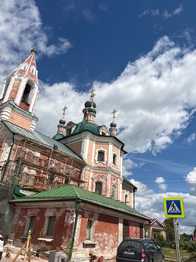 This image depicts an exterior view of a historic Russian Orthodox church under renovation. The church features traditional architecture with red brick walls, white decorative elements, and multiple green-domed cupolas topped with golden crosses. Scaffolding is visible along one side of the building, indicating ongoing repair work. The sky is a vibrant blue, with fluffy white clouds scattered across it, suggesting a bright, possibly daytime scene. A yellow pedestrian crossing sign is prominently displayed on a pole on the right side of the image. In the background, trees and other buildings are visible. A dark-colored car is parked near the church. The overall scene conveys a sense of historical preservation and community life. No people are clearly visible in the image.