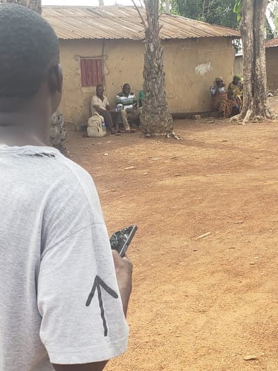 A medium shot captures a scene in the city of Kachia, Nigeria. In the foreground, the back of a person wearing a grey t-shirt with a black arrow drawn on the sleeve and a phone in their hand is visible. The t-shirt appears to have a tear near the shoulder. In the background, a simple, mud-brick building with a corrugated metal roof stands. Two men are seated on the ground in front of the building, one wearing a light-colored polo shirt and the other a striped green and white shirt. A light-colored backpack rests beside them. Further to the right, two women are also seated on the ground, one in a brown and white patterned skirt and the other in a green headscarf and yellow and brown patterned dress. They appear to be conversing. The ground is covered in red dirt. Trees are present, including a palm tree to the left and two large trees on the right.