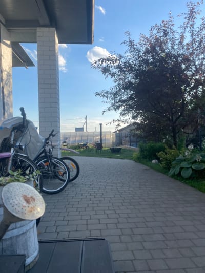 This media file depicts an outdoor scene on a sunny day, likely in the afternoon given the bright sky and shadows. The setting appears to be a residential backyard or patio area, possibly in a rural location given the presence of a greenhouse or polytunnel in the background and the overall rustic feel.

The primary subjects are two bicycles leaning against a white brick pillar. One bicycle is black with orange accents, and the other is silver with purple handlebars. Both appear to be used for personal transportation or recreation. In the foreground, part of a wooden barrel and a watering can with a perforated spout are visible, suggesting gardening activities.

The background features lush green foliage from trees and bushes, a paved walkway made of interlocking grey bricks, and a large, dark-colored structure that could be a shed or garage. Beyond the immediate yard, a greenhouse or polytunnel with translucent sheeting is visible, hinting at agricultural or horticultural pursuits in the area. A tall, slender structure, possibly a chimney or a weather monitoring pole, stands near the greenhouse.

The sky is a clear, vibrant blue with a few wispy white clouds. The sunlight casts distinct shadows, further indicating a bright, clear day. There is no visible text within the frame. The scene evokes a sense of everyday life, possibly a moment of pause after some outdoor activity. The location context provided, Мухавецкі сельскі Савет, Belarus, suggests this scene is situated in a Belarusian rural or semi-rural area.