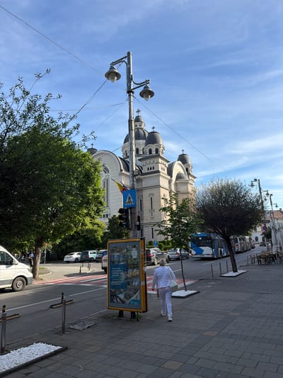 The image captures a street scene in Târgu Mureș, Romania. A significant portion of the image features the Orthodox Cathedral, exhibiting its distinct architectural design with domes. A person in white attire is walking on the sidewalk adjacent to the street. The street itself is lined with vehicles and trees, with a bus visible further down the road. A street light with spherical lamps stands prominently in the foreground. A yellow advertising display is present on the sidewalk. The sky above is partly clear, with some cloud cover.