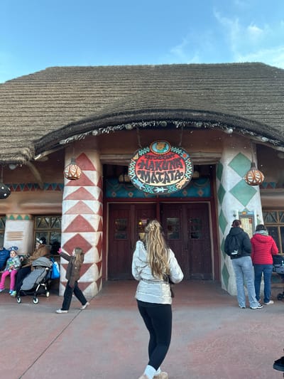 The image depicts an outdoor scene at the entrance of a themed building, characteristic of a theme park environment in Chessy, France (Disneyland Paris). The building features a prominent thatched roof and is adorned with patterned pillars and walls in earthy tones and geometric designs, suggesting an African or jungle theme.
A large, circular, and colorful sign hangs above the main entrance, clearly displaying the stylized text "HAKUNA MATATA." A small emblem, resembling a lion cub or warthog, is visible in the upper left portion of the sign.
Several individuals are present, indicating a bustling atmosphere. In the foreground, a woman with long, blonde, wavy hair, wearing a silver puffer jacket and black leggings, stands facing the building's double doors. To her right, two adults, one in a red puffer jacket and another in a black one, appear to be queuing or observing a display. Further to the right, a family group includes an adult pushing a black stroller that holds two small children, dressed warmly in a blue and pink jacket, respectively. A young girl in a brown coat walks nearby.
The scene portrays people visiting an attraction or restaurant within the park. The bright blue sky suggests it is daytime, and the warm clothing worn by many indicates cool weather. The overall setting and the "Hakuna Matata" text strongly imply a connection to Disney's "The Lion King" and Adventureland within Disneyland Paris.