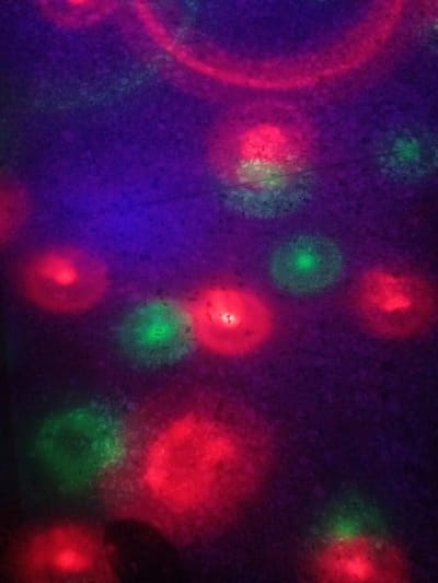 The image depicts a dark, textured surface illuminated by circular, diffused lights in red and green. The lights appear to be projected onto the surface, creating blurry halos. Some larger, fainter arcs of pink and red light are also visible, suggesting a more complex light source or multiple overlapping projections. The overall effect is abstract and atmospheric, with no discernible objects or people. The location in Sabon Gari, Nassarawa, Nigeria, is a residential area known for its vibrant markets and social life, but this image does not contain specific visual cues to identify it as such.