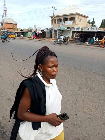 This image depicts a street scene in Tumfure, Nigeria. The central subject is a young Black woman with her hair in intricate braids, some of which are loose and trailing in the breeze. She is wearing a white top with a black garment draped over her shoulders and yellow pants. In her right hand, she holds a smartphone. Her expression is one of mild concern or focus.
The background reveals a typical street in a developing urban area. Several buildings line the road, some appearing to be shops with awnings. One building, taller than the others, has a light-colored facade and a prominent white roof. Signs are visible on some of the structures, including one for "Apple Drugs" and another with text in a local script, possibly indicating a food vendor such as "A.I.D SUYA SPOT." A large satellite dish is also visible.
Several vehicles are present on the road and roadside, including motorcycles, a yellow trishaw (auto-rickshaw), and a blue motorcycle with a rider. People are seen on and around the vehicles and in front of the shops. The time of day appears to be late morning or early afternoon, given the daylight and the angle of the sun, casting moderate shadows. The weather seems overcast, suggesting a possibly humid or temperate climate. The overall scene conveys a sense of everyday life and commerce on a Nigerian street.