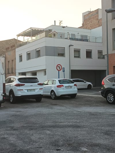 A white Hyundai Tucson SUV, license plate 7790 LFZ, is parked on an asphalt surface in front of a modern white building with multiple windows and a balcony on the second story. To the right of the Hyundai, a white Audi A3 hatchback, license plate 8105 HCB, is parked. A dark-colored SUV is partially visible on the far right. In the background, the white building has a darker gray band across its facade and a covered balcony with glass railings. A red and blue traffic sign with a red circle and a left-turning arrow with a red line through it, along with a blue "P" sign for parking, are visible on a pole. The sky is bright and appears to be either early morning or late afternoon. The scene is likely located in Tortosa, Spain, given the Spanish license plates and architectural style.