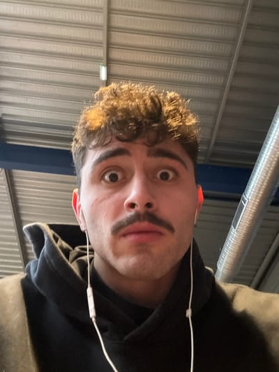 A young adult male, of light complexion with dark curly hair and a prominent dark mustache, stares directly at the camera with wide-open eyes, displaying a surprised expression. He is wearing a dark hooded sweatshirt and white wired earbuds are inserted into both ears, with the wires visible. The background reveals an indoor environment with a corrugated metal ceiling, a horizontal blue beam, and a silver cylindrical ventilation duct. A white rectangular fixture is visible on the ceiling. The scene is located in Chantepie, France.